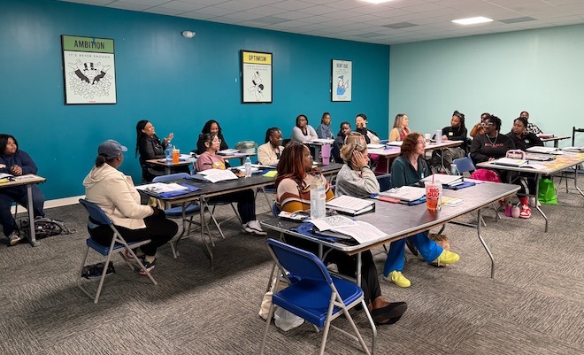 A classroom of women sit at shared folding tables before binders and papers. They are doulas in training in Richmond, VA.