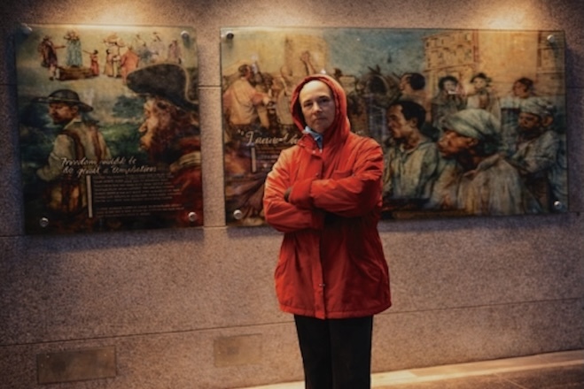 Lorene Cary, a Black woman in a hooded red windbreaker, stands in front of murals depicting slavery and the people President George Washington enslaved at the President's House exhibition at 6th and Market streets in 2010.