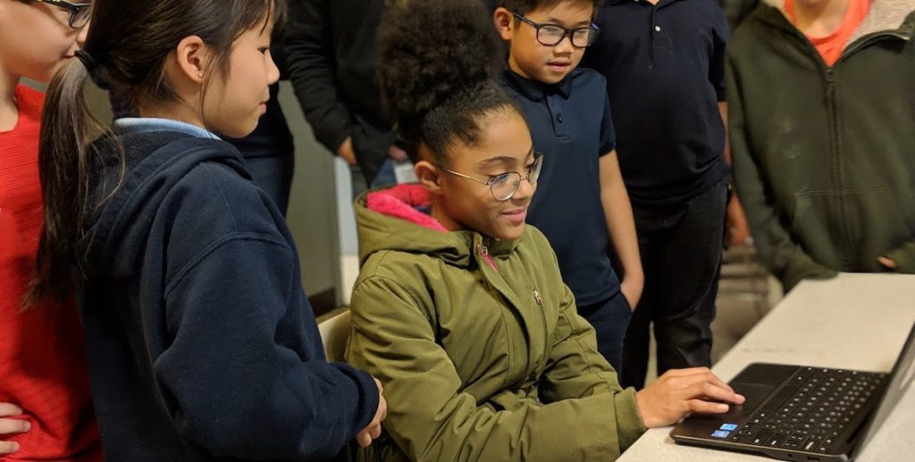 Children sit and stand around a computer.