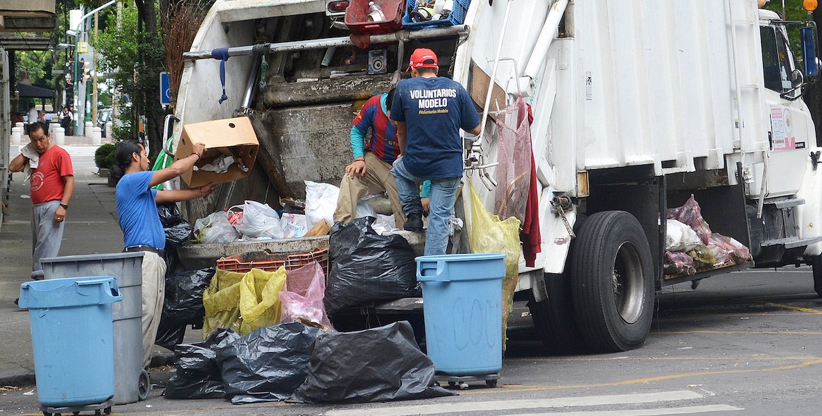 A large trash truck sits in the middle of the road while workers pile it with garbage.