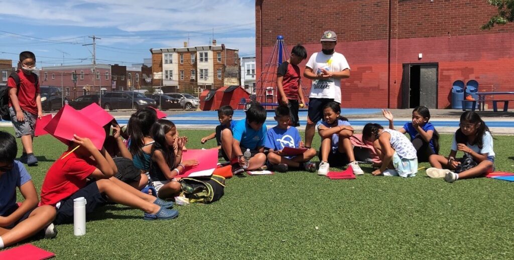 A group of young students and college-age Cosmic Writers instructors sit and stand on turf outside the Taggart School in Philadelphia.