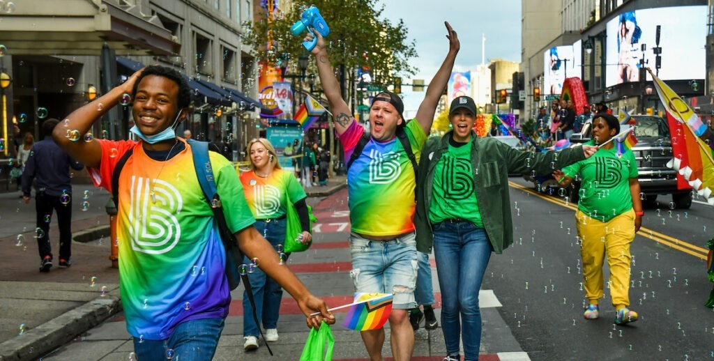 The National Coming Out Day Parade takes place every October in Philadelphia. In this photo, five people, three in rainbow tie-dye t-shirts, two in green t-shirts, most in jeans, walk jubilantly along Market Street amid bubbles.