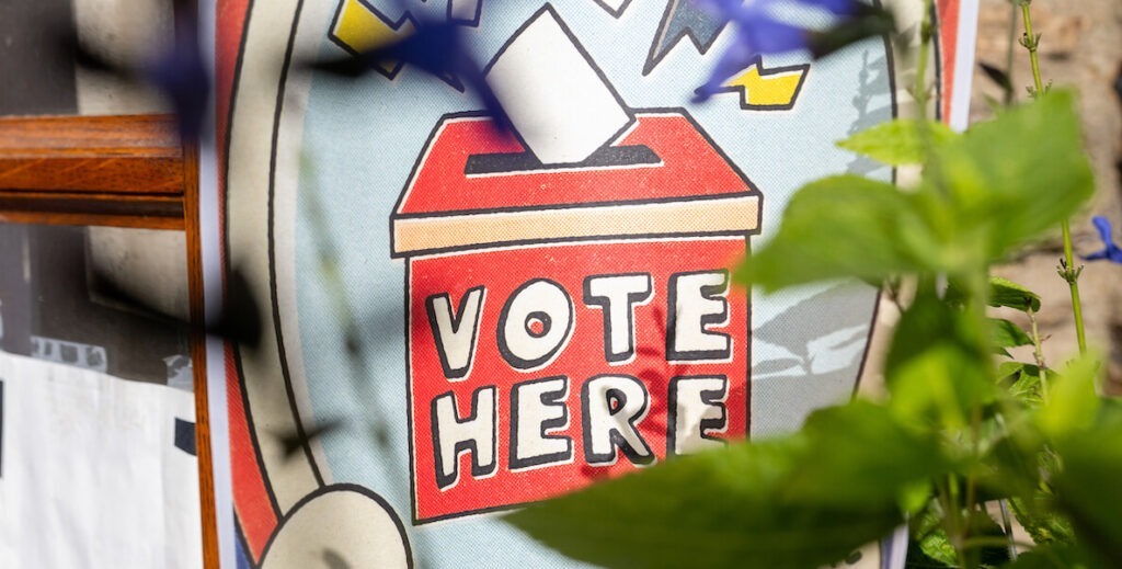 A outdoor poster or mural illustrates an old-fashioned ballot box with the words "VOTE HERE." There are lightning bolts around the box, and blank white ballots going into the box. In the foreground is a plant.