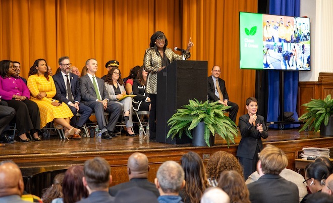 Mayor Cherelle Parker, a Black woman with glasses, thick straight black hair and bangs, wearing a gold jacket and shiny black blouse stands behind a podium giving a speech. Behind her are city leaders and officials. In the auditorium are heads looking toward her.