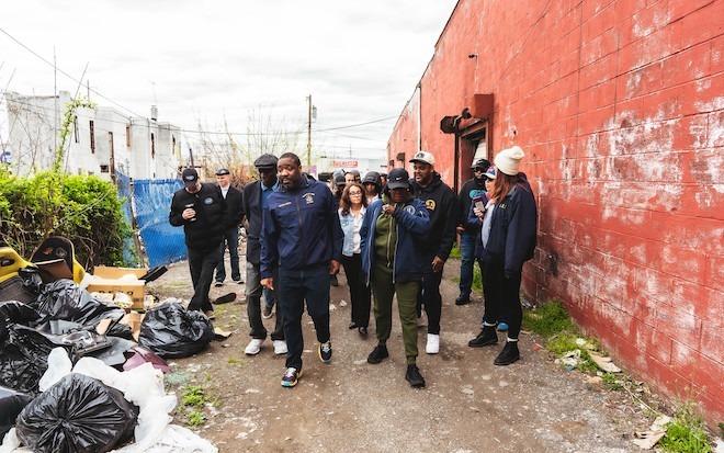 Left to right: Kenyatta Johnson, Mayor Parker, Carlton Williams (behind Parker), mostly Black and brown adults, tour an area between a faded building and piles of trash in Southwest Philadelphia. Photo by Chris Mansfield.