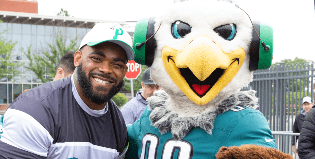 Eagles player Brandon Graham, wearing a white Phillies cap, smile alongside Eagles mascot Swoop, wearing a jersey and headphones, at the Eagles Autism Challenge.