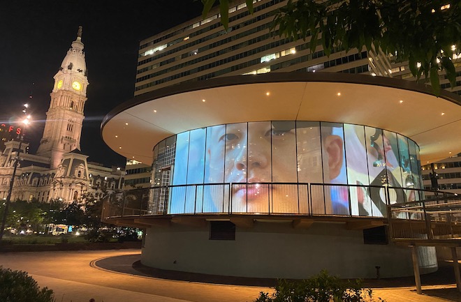 A projection of an Asian man's face on the circular visitors center in LOVE Park, across the street from Philadelphia City Hall.