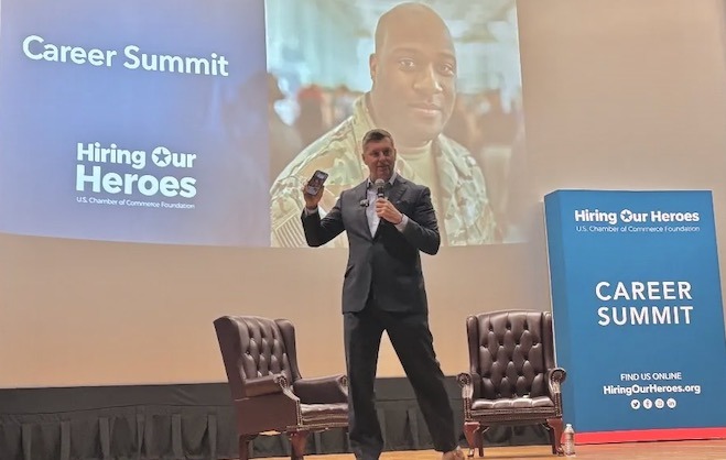 Patrick Murphy, a white man with short grey hair wearing a suit, stands on a stage in front of a screen with a photo of a man wearing Army fatigues. Murphy is speaking at the Hiring Our Heroes Career Summit in July