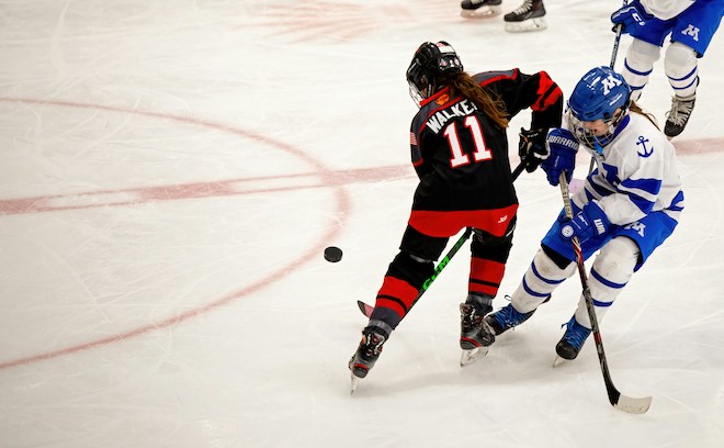 Two ice hockey players, girls with long hair, skate near the puck on a rink.