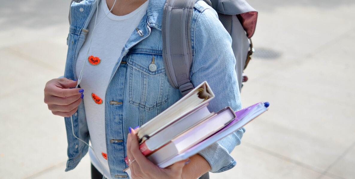 A student wearing a denim jackets and carrying an armful of books adjusts the volume on her Apple headphones.