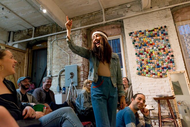 Artist Rose Luardo, wearing a long-hair wig, sweatband, blue jeans and a jeans jacket points in the air in front of a small crowd.