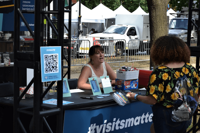 Kirsten Cornell, a White woman sits behind a table to represent the Pennsylvania Prison Society, and talks to a concertgoer with curly long hair and a sunflower blouse.