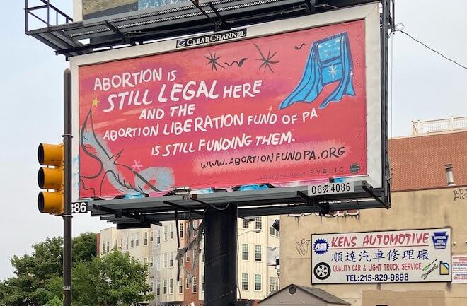 A pink billboard with an illustration of a blue window (created by Kah Yangni) reads: "Abortion is still legal here and the Abortion Liberation Fund of PA is still funding them. wwwabortionfundpa.org.