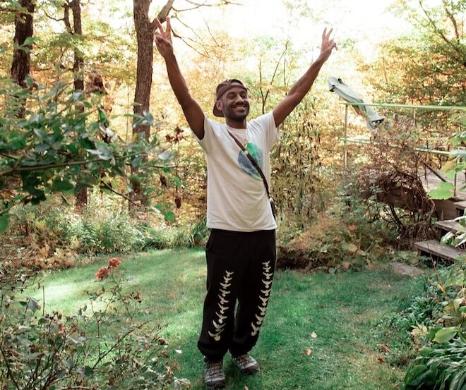 Saeed Ferguson, a Black man with a sparse beard, backwards baseball cap, t-shirt with an illustration of Earth, black pants with a white design holds up the peace sign on both hands and smiles in a wooded area.