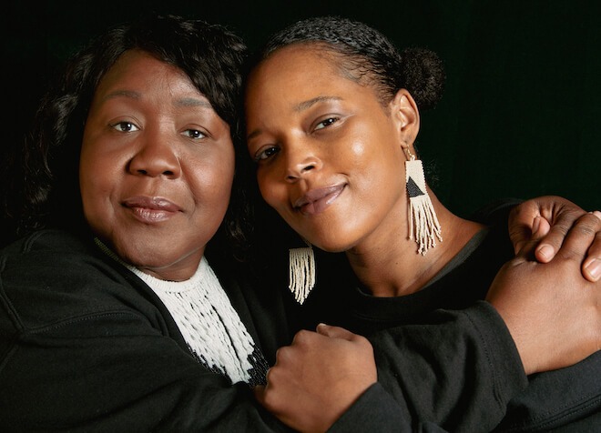 Poets Trapeta B. Mayson (left) and Yolanda Wisher, two African American women pose while hugging. They are wearing beaded jewelry — a necklace for Trapeta and dangling earrings for Yolanda — and wearing black shirts against a black background.