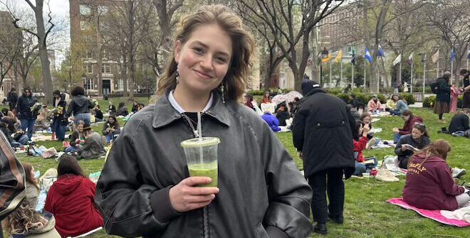 Madison Seidel, a white woman with wavy hair, stands in a dark jacket holding a cold matcha drink in a plastic cup, smiling among a crowd of women seated on blankets in Washington Square at a Dessert Before Dinner event.