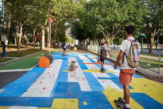 Skateboarders with backpacks, shorts, t-shirts ride along a painted pathway, strewn with leaves with trees on either side along the Benjamin Franklin Parkway in Philadelphia.