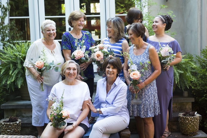 A group of women in their 30s through 70s, all wearing blue and violet, hold flowers and stand and sit outside a house window. They are volunteers for Forget Me Knot, an organization that repurposes wedding flowers for people in hospitals, nursing homes, shelters, and more.
