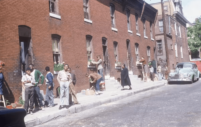 A group of Black residents on a street in Philadelphia are planting and maintaining window boxes on the side of a large brick building. A teal green 1950s automobile is parked at the end of the block