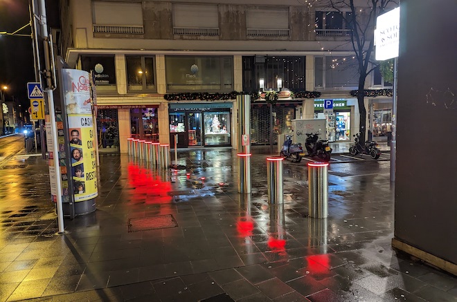 On a wet street, column-like silver retractable bollards with red lights around their top rim stand up to block vehicles from traveling along the street.