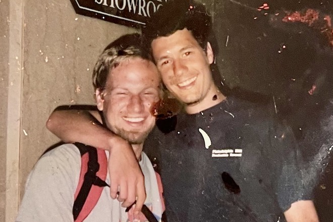 In an old photo, Kevin Hart, a white man with blonde short hair, a t-shirt, and backpack, stands next to Tim Judge, a taller White man with dark short hair in a dark t-shirt, with his arm around Kevin.