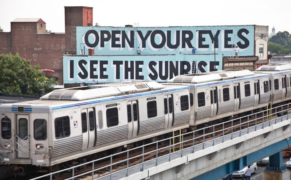 The El train in Philadelphia rides past one of Stephen ESPO Powers Love Letter murals that says, "Open Your Eyes I See the Sunrise." 