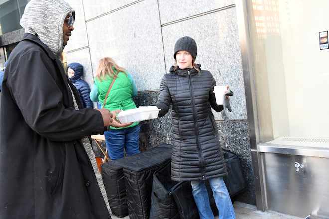 A woman in a winter coat hands a homeless man a container of food.