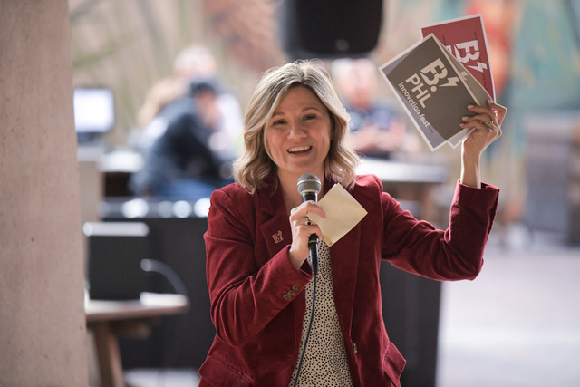 A woman in a maroon jacket and holding a microphone holds up cards that read B. PHL.