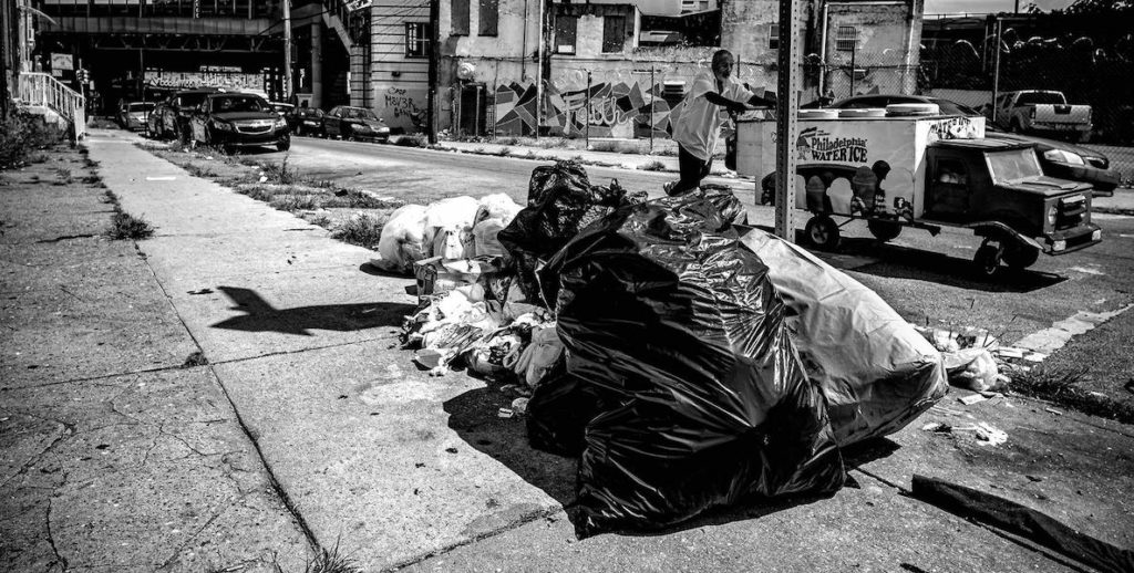 This photograph from Zamani Feelings shows several bags of trash lying on the ground as a man strolls by pushing a mini water ice truck.