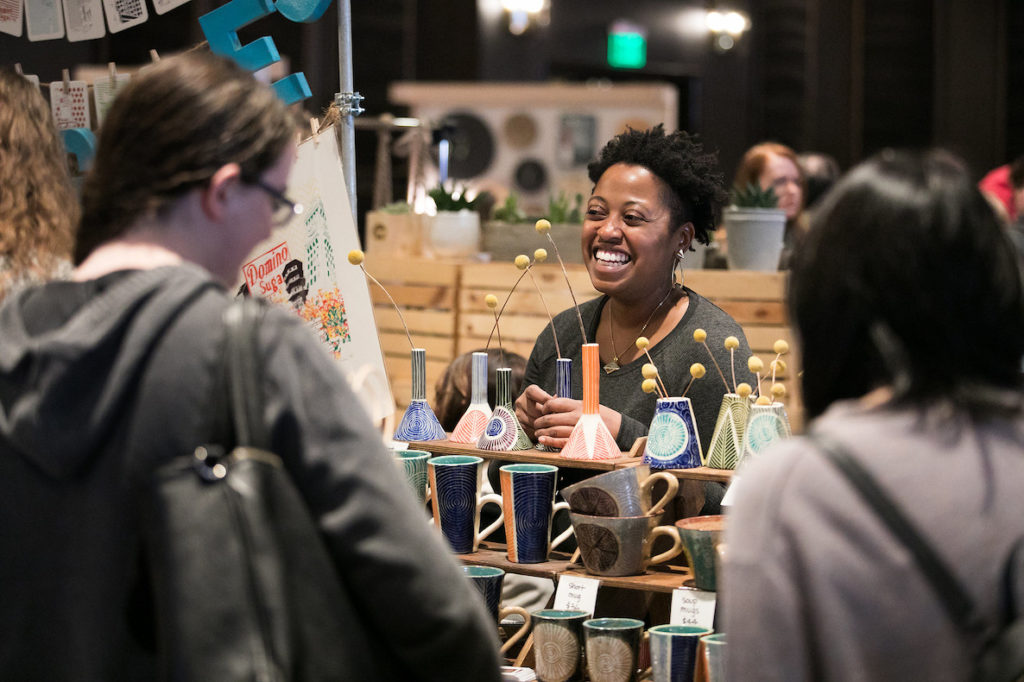 A woman smiles at a customer from behind her booth at the annual Holiday Art Star Craft Bazaar