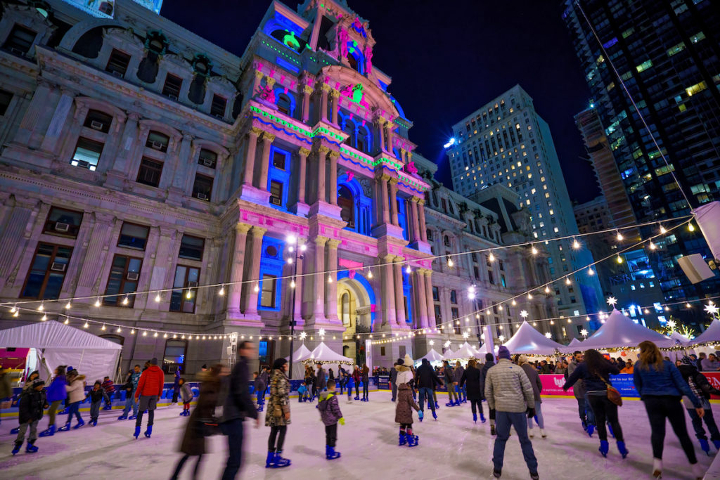 A crowd of people twirl around the ice skating rink at City Hall, with City Hall all aglow in the background.