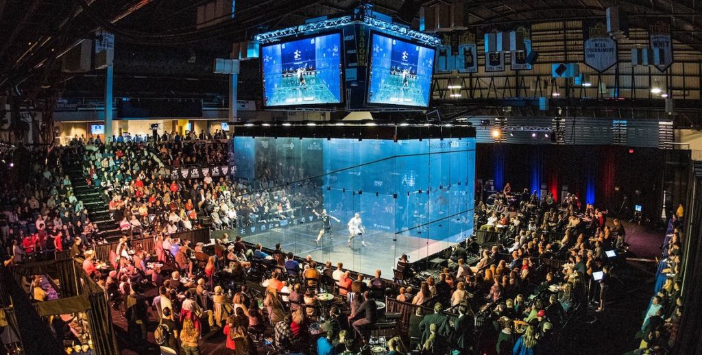 A game of squash takes place in a glass cage, while onlookers stand around and watch.