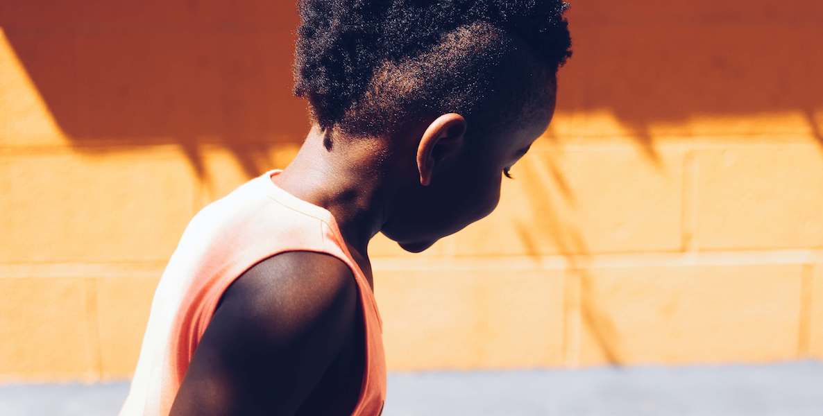 A young black girl in a tank top looks away from the camera. There's a bright orange wall in the background.