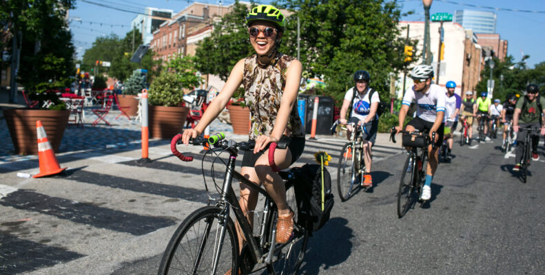 A woman rides through South Philadelphia on her bicycle, smiling big with a helmet on.