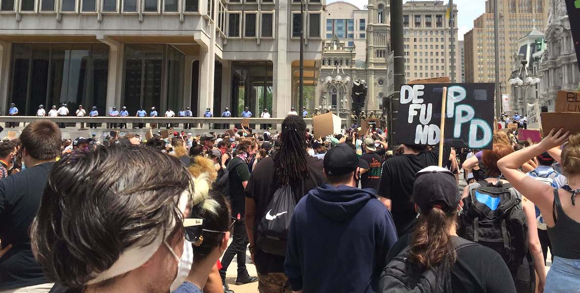 Citizens gather for a defund police rally and protest in Philadelphia, while the Philadelphia police line up and look on in the background.
