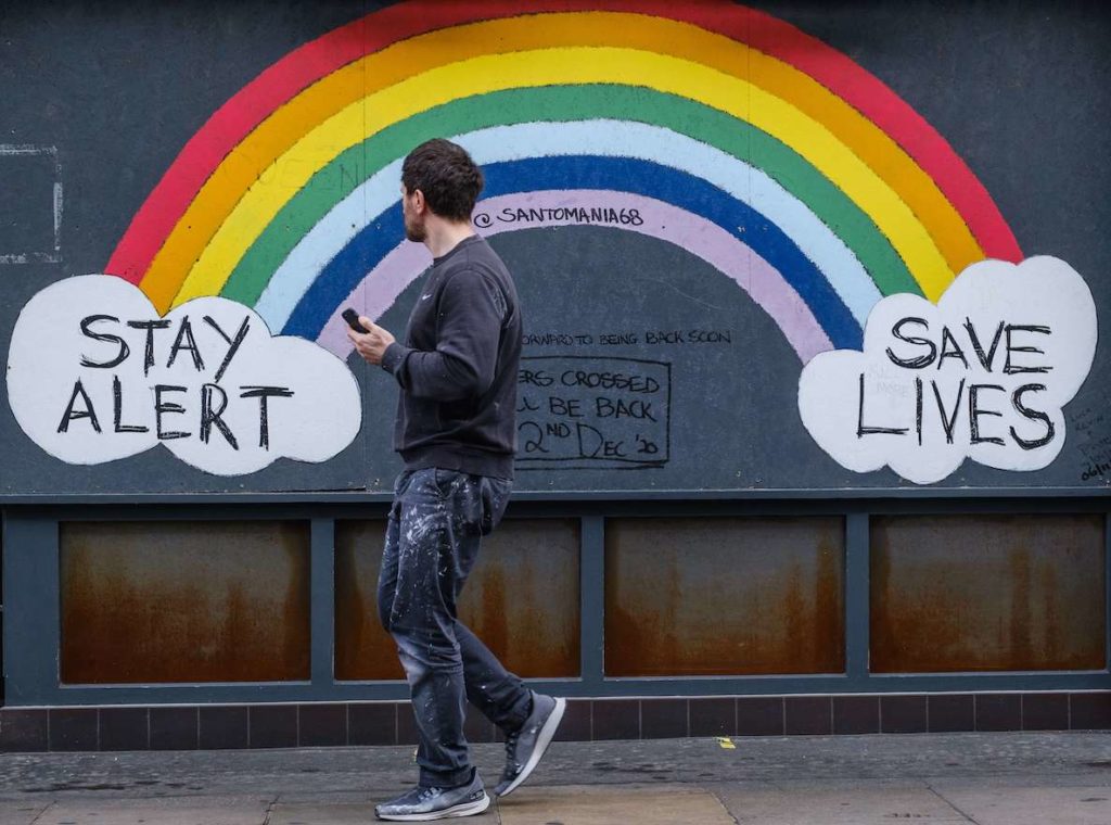 A man walks past graffiti on the wall: a rainbow with two clouds on each end that say "Stay Alert, Save Lives."