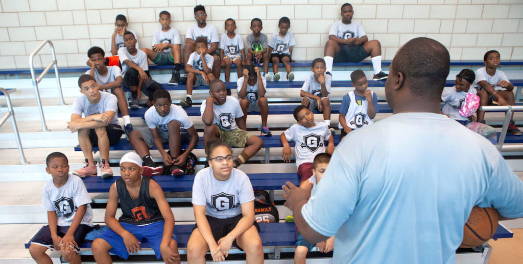 A coach speaks to players sitting in bleachers and participating in a Give and Go Athletics sporting event.