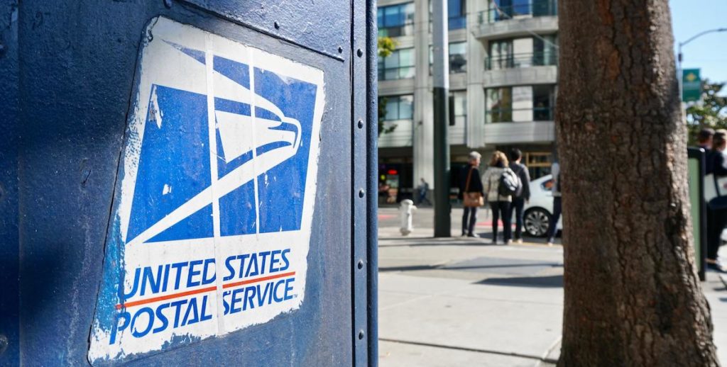 A United States Postal Service logo is emblazoned on a blue mailbox, in the background people stand at a street corner waiting for a cab.