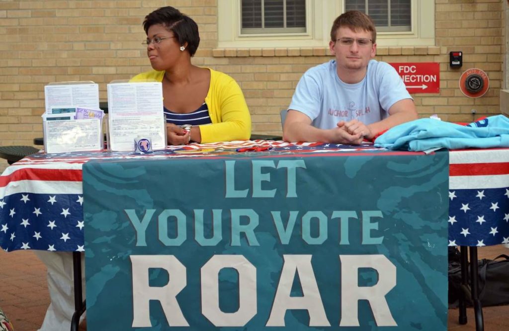 A man and woman sit at a voter registration table that has a sign hanging on it that reads "Let Your Vote Roar"