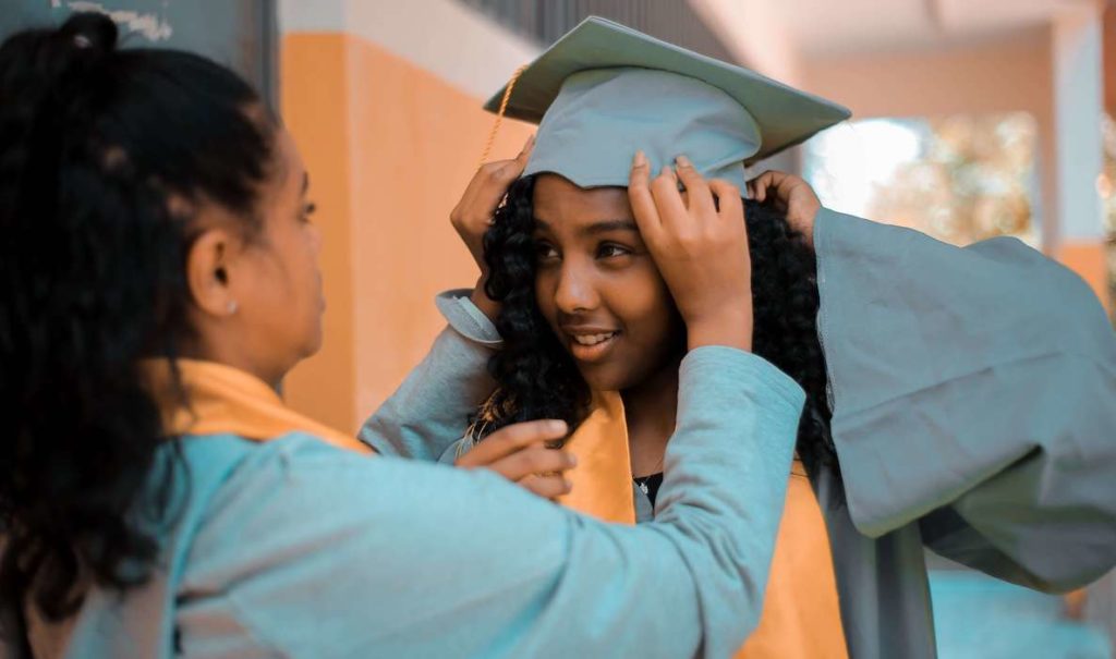 Two women help each other put on their graduation hats before the ceremony