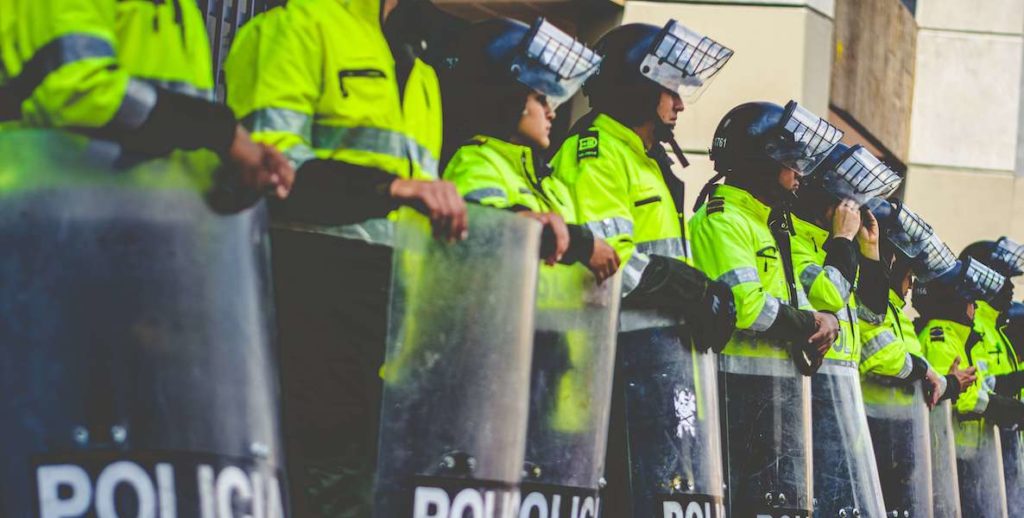 Cops wearing a neon yellow color line up holding shields and wearing masks