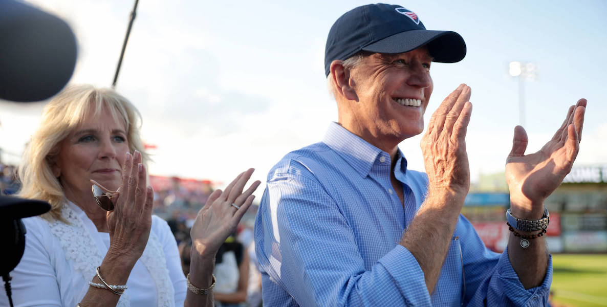 President-elect Joe Biden, wearing a cap, claps at a baseball game, with First Lady-elect Jill Biden standing behind him