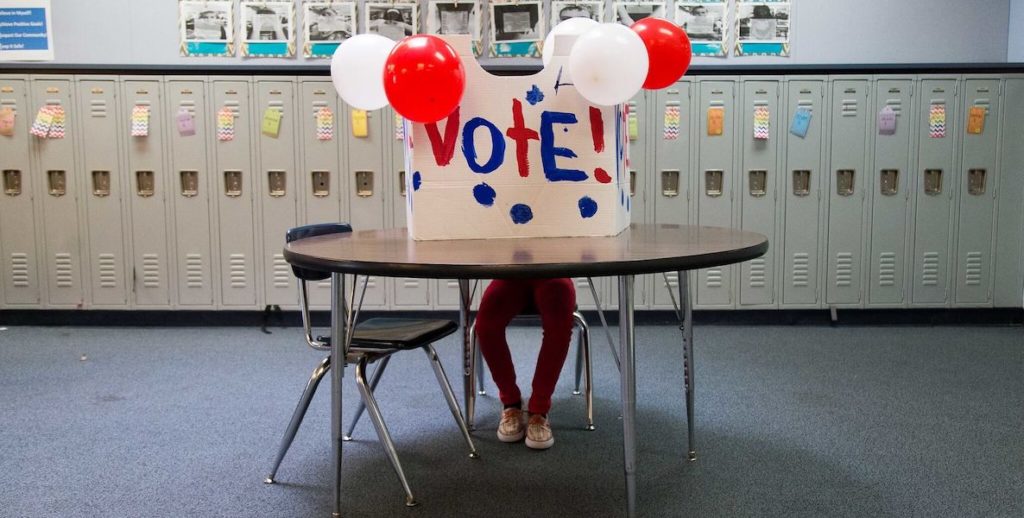 Someone sits behind a hand-painted vote sign with balloons attached.