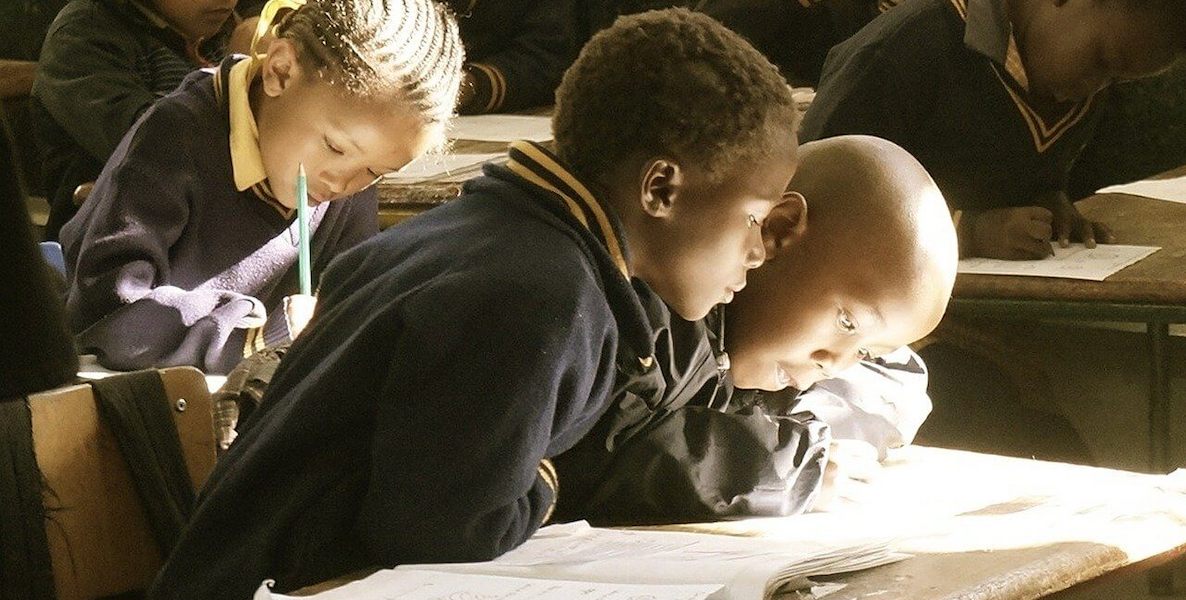 Two young black students huddle together doing their school work as a ray of light shines through the window onto their desk.
