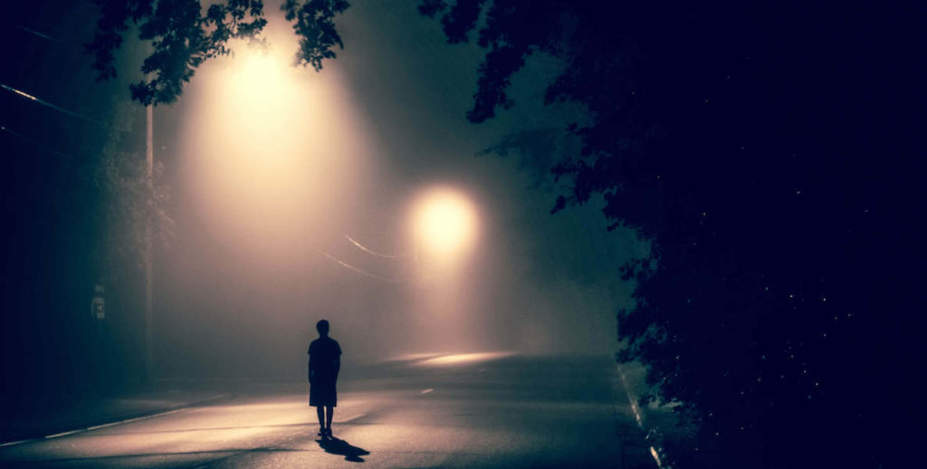 A person walks alone at night along a street lit by street lamps
