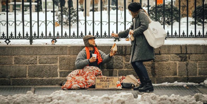 A woman wearing a mask donates a sandwich to a man on the street on a cold, snowy day.