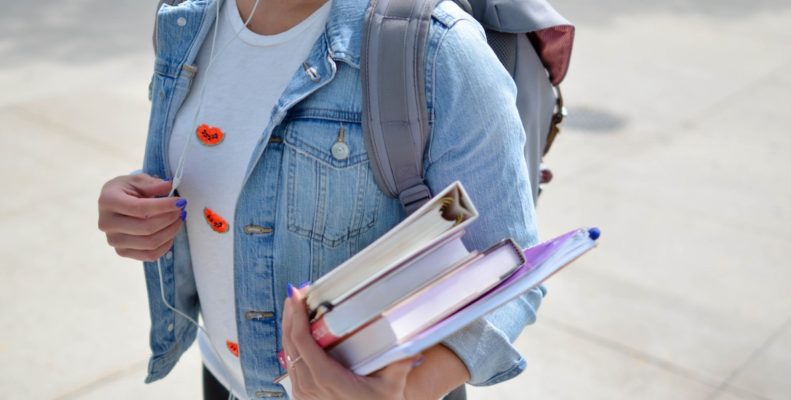 A student wearing a denim jackets and carrying an armful of books adjusts the volume on her Apple headphones.