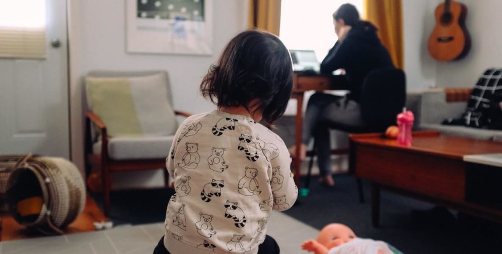 A working mother bangs away on her computer while her daughter plays in the background. Universal child care would be nice for her and so many other women—and men—like her.