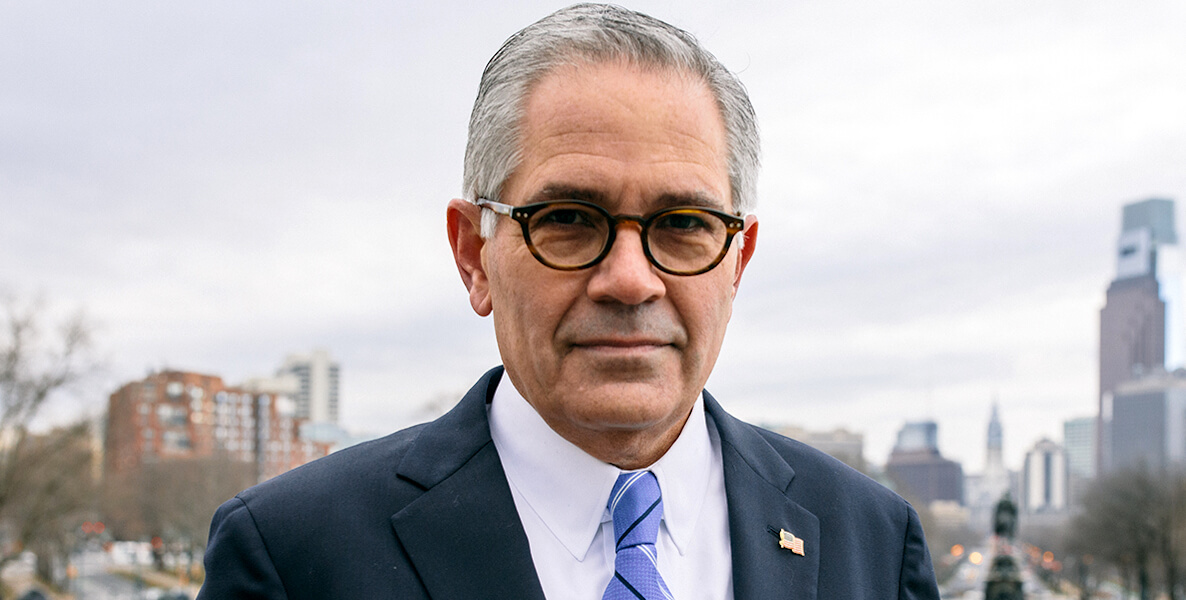 Philadelphia District Attorney Larry Krasner stands on the steps of the Philadelphia Museum of Art, with the city of Philadelphia skyline in the background.