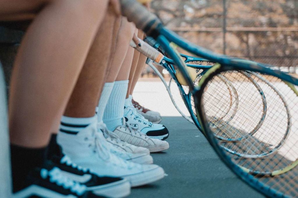 Kids holding tennis rackets and wearing tennis shoes await their turn on the tennis court
